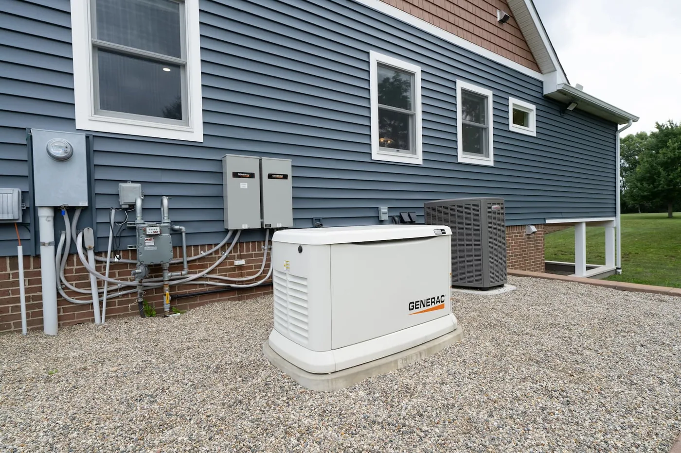A Generac standby generator installed on its pad beside a Fishers home, with two transfer switches, the gas line, and the electric meter on the wall behind it.